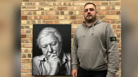 John Donaldson next to his painting of Sir David Attenborough. John has short dark hair and facial hair and is wearing a grey hoodie. He is stood next to the black and white portrait of Sir David Attenborough. In the painting, he has his hand on his chin, slightly covering his mouth. He is looking down, deep in thought. 