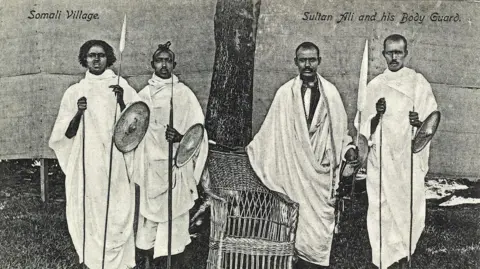 A historic black and white photograph of four men in white robes holding spears and shields