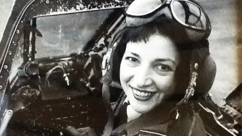 Candy Adkins Black and white photo of a smiling Jackie Moggridge sitting in an aircraft cockpit. The canopy is open and she is turning back to look at the camera behind her. She is wearing a headset and a pair of flying goggles on top of her head.