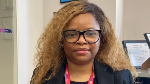 A woman with brown/blonde curly hair and black-rimmed glasses looks at the camera. She is wearing a black top under a black jacket and has a pink lanyard around her neck.