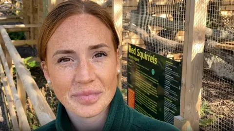 Luke Deal/BBC A photo of Emily Quantrill. She is facing the camera and smiling. She is standing in front of the red squirrel enclosure. She has ginger hair which is tied back and she is wearing a green jumper and a set of pearl earrings. 