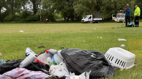 Piles of rubbish in ripped bin bags on green grass with council workers and vehicles in the background