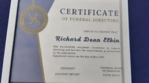 CPS A cropped framed image of a faked certificate of funeral directing with the logo of National Association of Funeral Directors and Richard Elkin's name on it. The wall it's hanging on is purple.