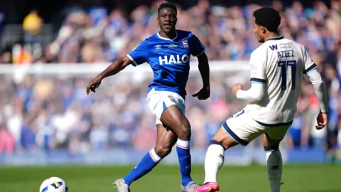 PA Media Two football players mid-tackle on a football pitch. One is wearing blue and white kit and the other is wearing white kit. 