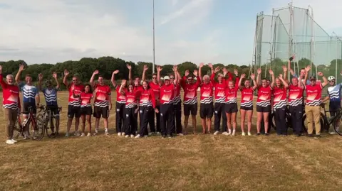 Team members in a line, wearing red Isle of Wight tops, waving to the camera