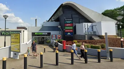 Membury Services - a building with a domed roof with bollards in front of it and people walking in and out. The sign above the door says, "Welcome Break" and there is a board outside to the left listing all the shops inside. There is also a red postbox.