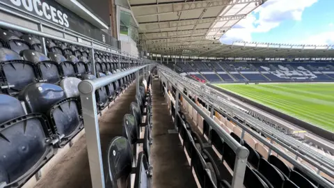 The view along a row of safe standing seats at a sports stadium. Round steel barriers extend into the distance, each in front of a row of black plastic seats. The grass pitch is visible to the right.