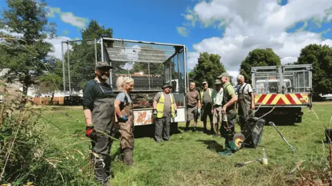 Trout in the Trym A group of volunteers standing in a field by some council rubbish vans. The are wearing protective dark green clothing and a wheelbarrow in an upright position is near the front of the picture. 