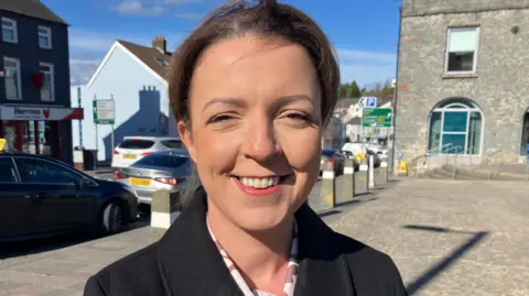 BBC Liz Kimmins smiles at the camera. She has brown hair and wears a back coat. Cars and buildings in Ballynahinch are behind her.