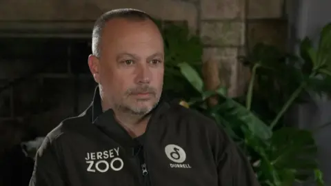 Mark Habben is sitting in front of a granite fireplace with a green cheese plant to the right side of the background. He is a white man with short grey hair and a grey beard, wearing a Jersey Zoo-branded brown jacket. He has a serious expression on his face.