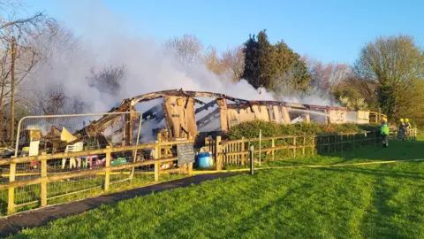 A collapsed burnt out single storey building with smoke rising. The building is in a park with a wooden fence around it and firefighters can be seen spraying water on to the building from behind the fence on the right of the picture.
