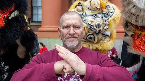 Tommy Wong Stephen Ornellas with short grey hair and beard wearing a burgundy top with the Liverpool Hung Gar Kung Fu School logo does a martial arts pose as he stands in front of Chinese lions. 