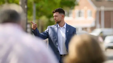 Getty Images A man in his thirties wearing a blue suit jacket and white shirt. He is stood in a public street and pointing to something.