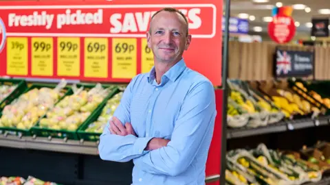 Aldi Giles Hurley, Aldi UK chief executive, poses with his arms folded in front of the fruit and veg aisle in an Aldi supermarket