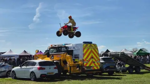 Sam Coulter A man on a quad bike in mid-flight while jumping over two cars, a digger and a police van. It is a sunny day at a show and there are marquees in the background. The stuntman is wearing a yellow suit, red and yellow helmet and his quad bike is  yellow and red. A ramp is propped up before the first car.