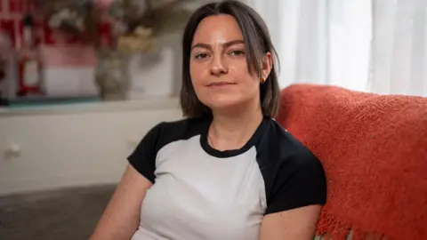 Lucy Asson has shoulder-length dark hair and is wearing a white t-shirt with black shoulders
