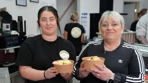Joanne and Charlie  Bruford are standing behind the counter of their new pasta and salad bar holiday a salad box each. Charlie has dark hair toed off her face, Joanne has blonde hair with a fringe tied off her face. Both are wearing black.  