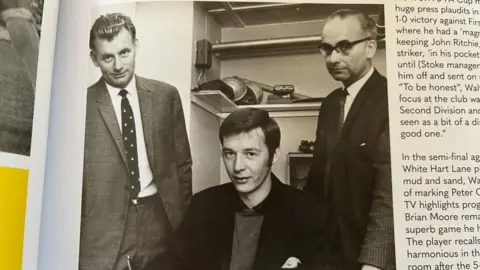 Nicola Haseler/BBC A black and white photo of Walter Lees as a young man sitting at a desk about to sign a contract. He is flanked by two other men in suits who were the managed and secretary at the time. 