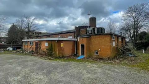 A former social club site, with bushes next to the brick building. There is grass to the right hand side of it and trees are in the background to the left and right. 