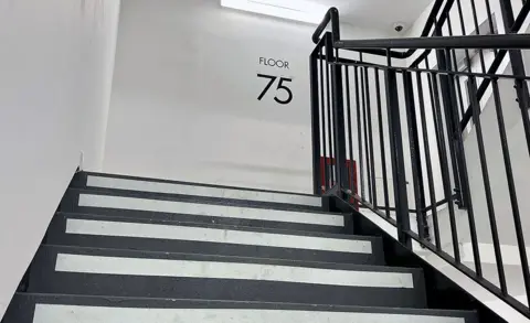 A presumption    of the staircase astatine  the Landmark Pinnacle gathering  with a motion   connected  the partition  astatine  the apical  of the stairs saying 'floor 75', the stairs are achromatic  and achromatic  with metallic bannisters and the walls are white.