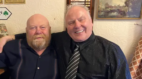 Two men are standing side by side with thier arms around each other. The man on the left is wearing a dark blue shirt with a pink stripe on either side of his shoulders and the man on the left is wearing a black shirt with glitter strips and a black and white tie. They are stood by a wall in a pub