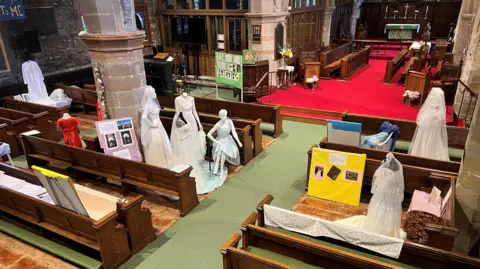 A church interior, seen from a height, with six standing mannequins clothed in long white wedding dresses, three with veils, and one seated one displayed between the pews. A smaller mannequin wears a long red dress, and there are a number of poster boards with photos.