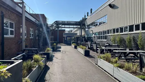 Emma Baugh/BBC News Two industrial buildings with a walkway through the centre with seating and planters on both sides with stairs to upper levels with workers enjoying the sunshine
