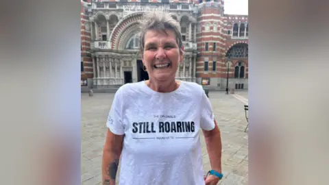 Lynda Hale smiling for the camera. She is wearing a white T-shirt with inscription "The Originals STILL ROARING squad of '72"