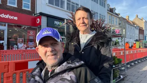 Andrew Turner/BBC Jane Cole, on the right, with dark hair, wearing a light top and black coat, with her partner David Durrant, in a black and grey jacket and blue baseball cap. He is bearded and is using a wheelchair. They are pictured near some shops and a market flower stand, and some orange roadworks barriers