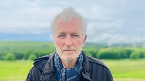 Ian Baird, a man with grey hair and grey stubble, looking at the camera, he is wearing a dark jacket and a blue scarf, with a grassy landscape behind him.