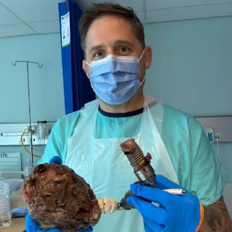 Rhys Thomas Rhys in hospital holding his heart which is attached to a pipe. He is wearing blue hospital pyjamas, a platic clear apron, blue plastic gloves and a blue surgical mask.