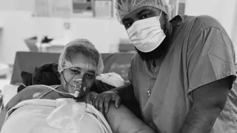 Grace Victory Grace and her partner Lee in medical gowns, Grace is wearing a breathing mask and Lee is wearing a paper mask