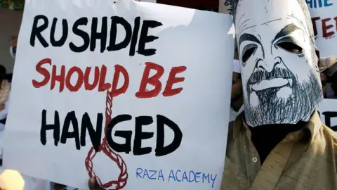 On January 12, 2004, during a protest in Mumbai, an Indian Muslim wore a mask of Indian writer Salman Rushdie and held a placard condemning Rushdie. Dee appeared at protests in the city, offering a reward of 100,000 rupees ($2,199) to anyone who discredited Rushdie, who allegedly wrote against Islam's Holy Prophet in his book 