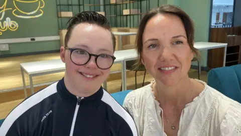 Lara King A smiling teenage boy in a blue and white tracksuit sitting next to his mother, a woman with brown hair and a cream blouse.