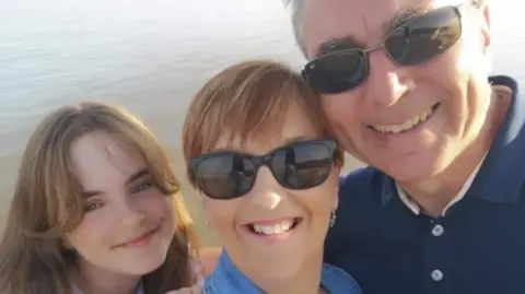 Family photo A selfie of Marianne with her husband and young daughter. They are by the sea and she and her husband are wearing sunglasses.