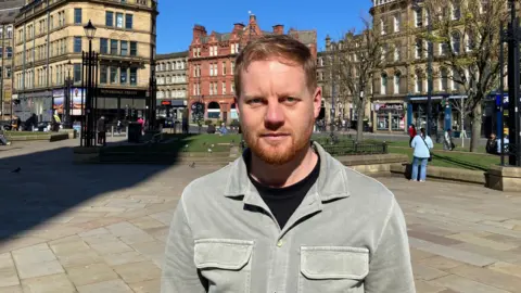 A man with a ginger beard and strawberry blond hair wearing a light grey/green shirt and black T-shirt standing in Bradford city centre 