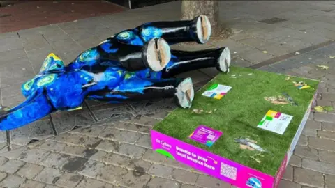 St Giles Hospice A blue Vincent Van Gogh Starry Night themed elephant lies on its side on the ground, next to the green fake grass topped plinth it was originally on