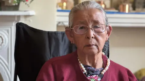 Wearing a bright red card cardigan, a string of pearls around her neck Devisara Rana is sitting in her bedroom on a black camping chair. Wearing glasses her expression reflects the fact she's lost both her husband and son in recent years. 