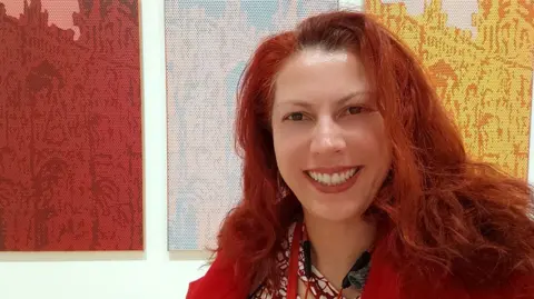 Helena Diez Helena Diez has red hair and is standing in front of three pictures - she has a red and white shirt on and is wearing a landyard