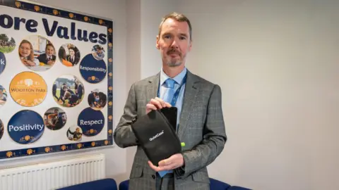 Northamptonshire Police, Fire and Crime Commissioner A man in a grey striped suit and blue shirt and tie holds a black pouch with a zip on the side. "Quiet Case" is written in white lettering on the side. 