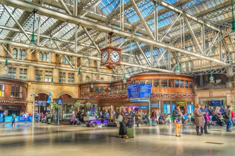 Alli McMahon A colourful scene of people waiting in the station for trains. There is a clock hanging from the roof and brown kiosks in the background.