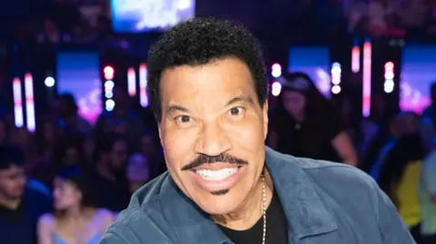 A black man with short, curly, black hair and facial hair is smiling toward the camera. He is wearing a a blue suede jacket with a black t-shirt underneath and a silver chain around his neck. 