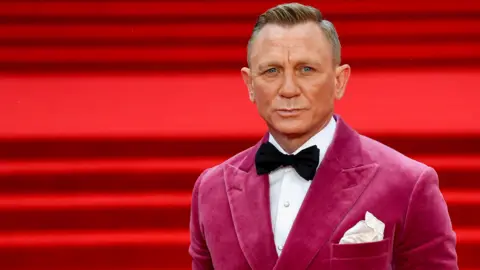 Reuters Daniel Craig standing on a red carpet for the world premiere of No Time To Die. He is wearing a black bow tie and a dark pink dinner jacket