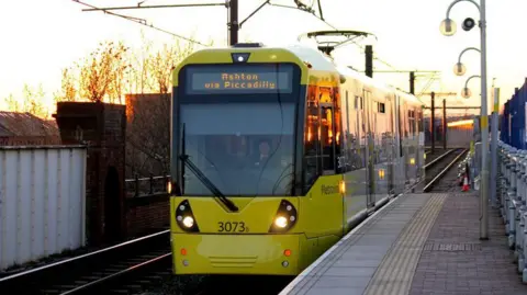 A yellow and grey tram on a track at a platform outside. The sun is setting in the background. 
