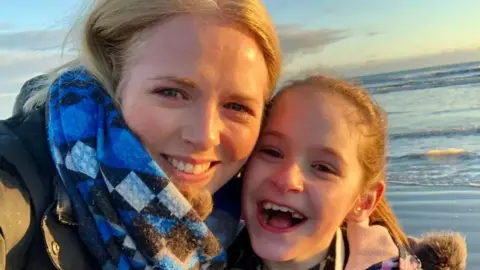 Helen Husbands A blonde woman in a blue scarf smiling and looking at the camera while holding her head close to a young girl who is also blonde and smiling with her mouth open.