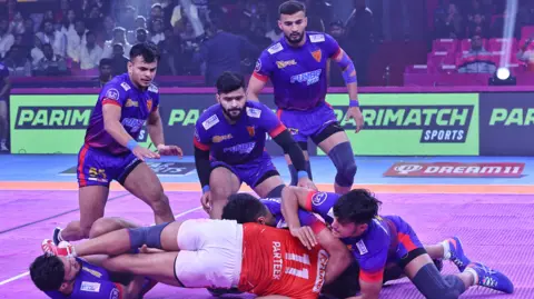 Getty Images One man in red, being tackled by several men dressing in a purple kit with a crowd watching on in an indoor arena