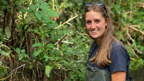 Fiona Irving / BBC A woman with long brown hair and sunglasses on her head smiles at the camera. She is wearing green waders and a blue t-shirt. She is surrounded by green vegetation.