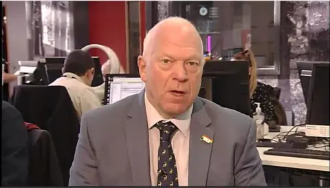 Stephen Giles-Medhurst wearing a grey jacket with a white shirt and dark grey tie which has a small white logo appearing several times. There is a media newsroom behind him.