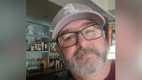 Supplied A selfie-style image of Richard Andrews. He is wearing glasses and a baseball cap and standing in front of a bar. He is looking into the camera. 