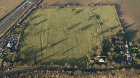 Historic England Archive Aerial view of a green field surrounded by hedges. Beneath the ground lies the remains of the Norman Cross Camp.